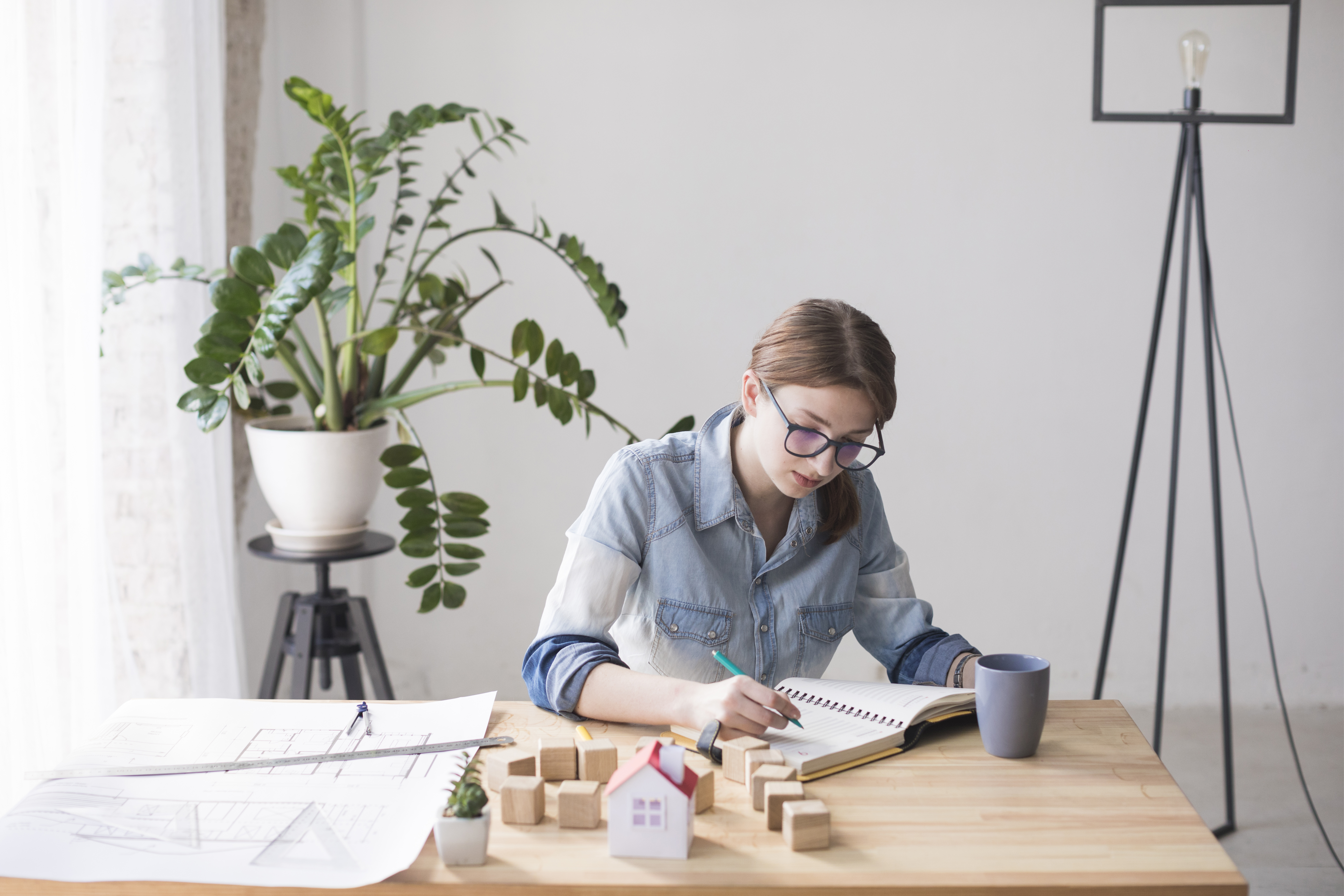 Mujer joven sentada en una mesa de madera, rodeada de bloques en forma de casas y una maqueta de vivienda, representando seguros inmobiliarios.